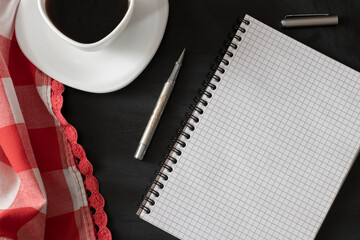 White cup of strong black coffee with metal pen, notepad and red-white cloth on black surface close up