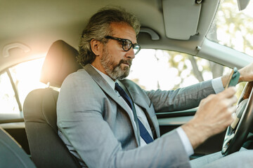 Close up of smiling old businessman using smart phone while sitting in his car. Focus on phone.