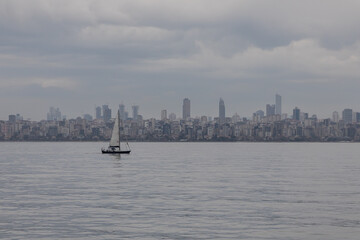 A small lonely boat is floating under the sail in the wide sea space against the backdrop of the gloomy faded city line of Istanbul along the coast. Cloudy dull day at the sea.