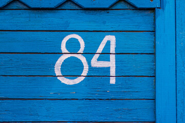 House number 84. The numbers are painted in white paint using a stencil on a wooden blue wall made of dry old planks. The wall of the village house