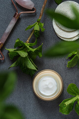 Facial roller, gua sha,moisturizing cream and green leaves. Natural cosmetics and facial tools on dark marble background.Top view. Flat lay composition.