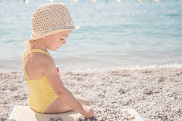 Cute little girl is playing with pebbles on the beautiful beach. Child relaxing on beach resort.