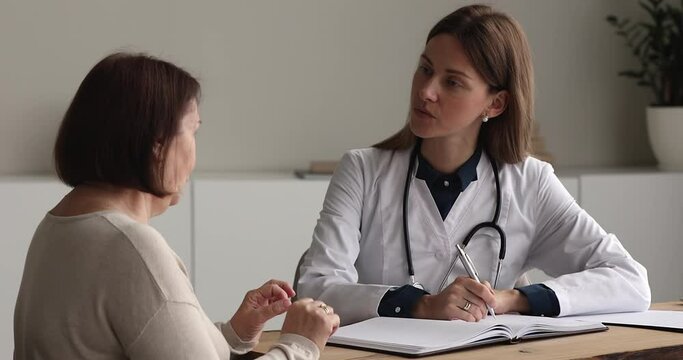Woman doctor in white coat sit at desk writes in journal complaints and health concerns of older patient during visit in clinic. Medicine, medicare for old citizen, professional medical aid concept