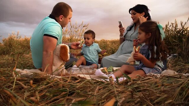 A family picnic on nature in a summer field at sunset in slowmo. A caucasian couple with two children having a rest in a meadow. Father is feeding children and mother is taking pictures on her mobile.