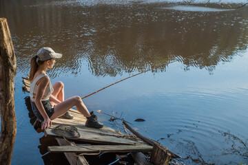 A girl with a fishing rod on a wooden pier is fishing in a forest lake.
