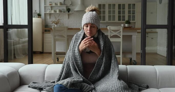 Young woman wear hat wrapped in knitted plaid sit alone shivering from cold on sofa in unheated apartment without central heating due debt. Unhealthy female feeling discomfort try to warming up indoor
