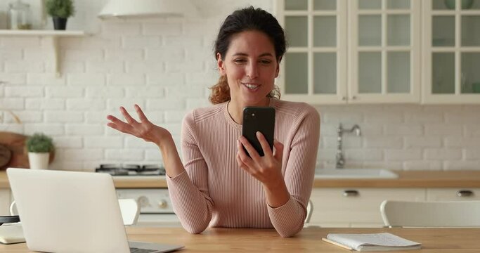 Woman spend time in kitchen use smartphone wave hand greeting relatives or friend virtually, enjoy distant web chat. Modern technology and video conference application usage, video call event concept