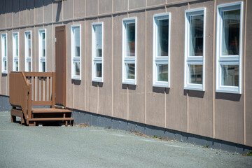 The exterior of a commercial building, school, with multiple tall narrow windows with white trim. The wall is tan color wood. There's a single metal brown door with a small patio and step. 