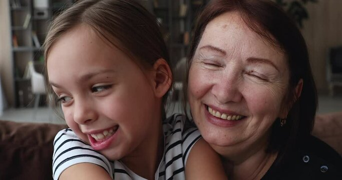 Close up happy cheerful faces of older 60s granny and little granddaughter sit on sofa look at camera making selfie or videocall, recording video together, capture moments. Family fun and love concept