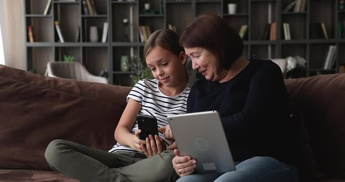 Older granny and little granddaughter holding tablet and smartphone while resting together on sofa at home. Multi-generational family discuss new video game, enjoy free time using modern tech concept