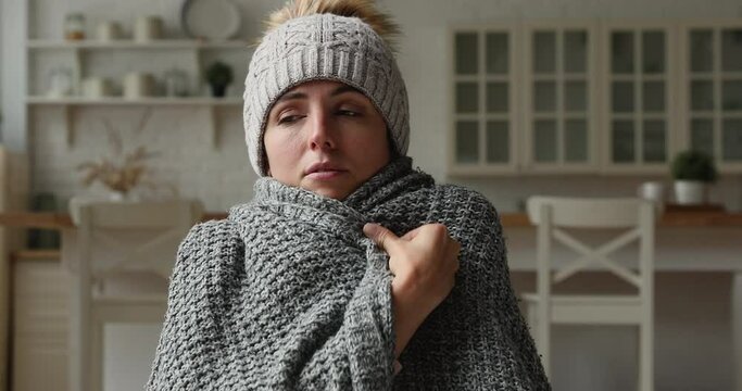 Young millennial woman try to warm up wear grey knitted bobble hat, scarf, wrapped in warm plaid, shivers from cold sitting alone at home, close up. Radiator dont warmup, central heating debt concept