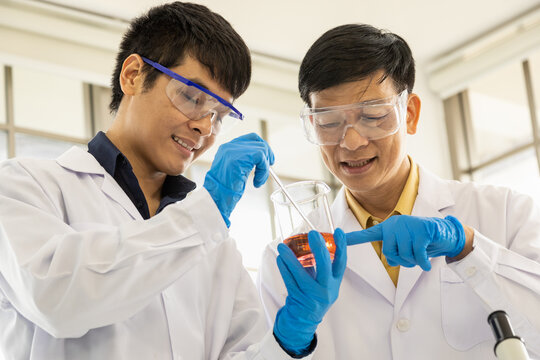 Professional senior doctor scientist teach and guide young scientist looking to beaker with orange liquid substance in working laboratory.