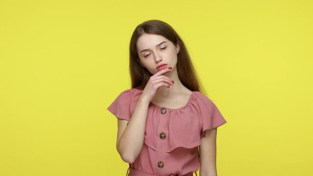 Thoughtful girl with brown hair in elegant dress, keeping finger on her cheek, looking aside with pensive expression, finding solution for doubting question. Indoor shot isolated on yellow background.