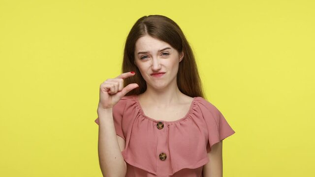 Cute female looking at camera with disappointed pitiful expression and showing a little bit gesture, feeling skeptical about small size, wearing pink dress. Indoor shot isolated on yellow background.