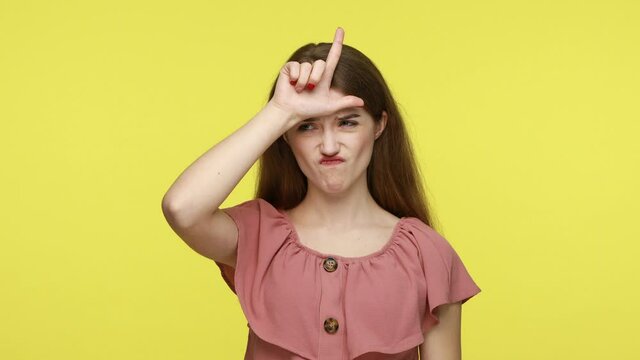 I lose! My defeat! Unlucky sad woman in dress making loser gesture, L sign on forehead and sighing with sadness, feeling desperate about unsuccess. Indoor studio shot isolated on yellow background.