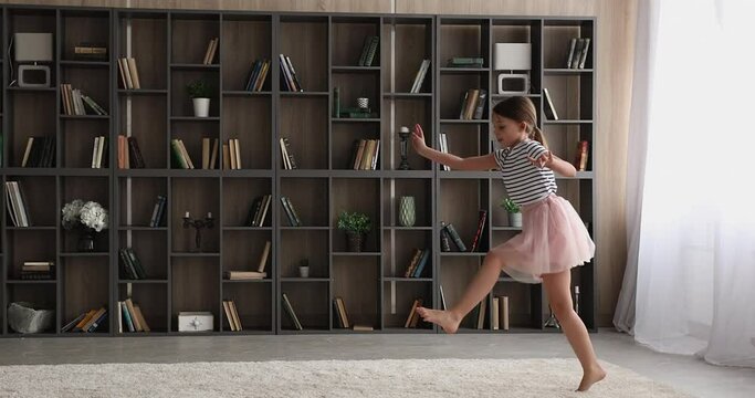 Lively vivacious girl wear fluffy festive skirt dancing barefoot on carpet in modern living room. Little child listens funky music jumping have fun alone at home. Hobby, dance school for kids concept
