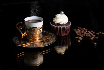 Steaming golden coffee cup, homemade cupcake and coffee beans on black background with reflection