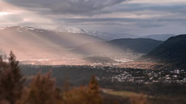 Sun rays piercing the clouds and shining over the Mo i Rana town in Northern Norway. The thick cover of the blue-pink clouds hanging in the sky. Mountains covered with dense forest surround the valley