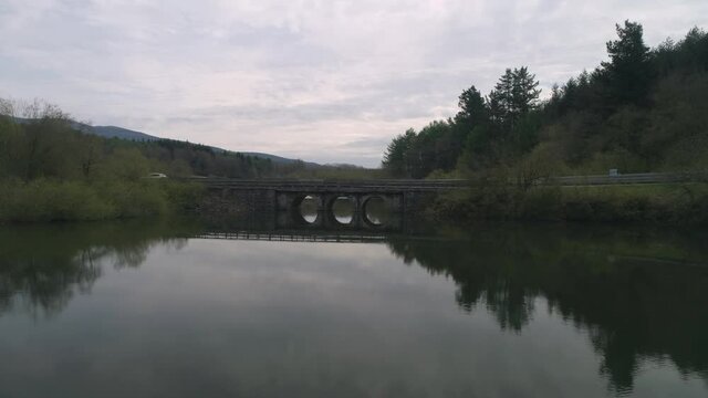 Car driving on the stone bridge over a still lake with its reflection in the water - aerial sideways