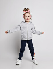 Cute smiling active kid girl in gray hoodie with zipper is dancing with arms outstretched, jumping...