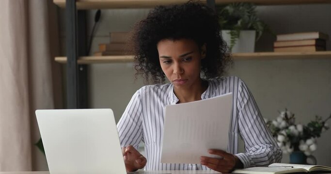 Serious young African ethnicity woman employee sit at workplace desk holding document sheets, do paperwork learn information, analyzing correspondence, make financial papers research, workflow concept