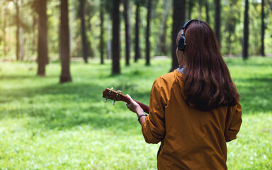 Rear view image of a woman enjoy listening to music with headphone while playing ukulele in the park