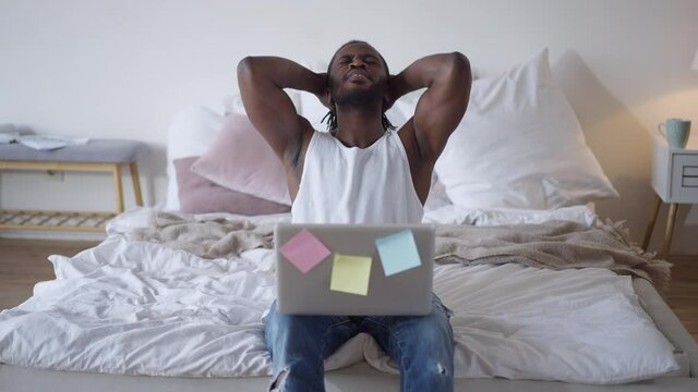 Front view of African American young man leaning back on bed as laptop falling and guy catching device. Cheerful tired freelancer working online from home on lockdown isolation