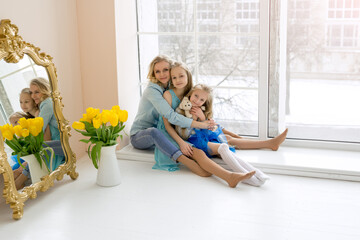 Mom hugs her daughters sitting at the window. A bouquet of yellow tulips in a vase stands by the window. Happy family. Flowers from daughters for mom on Mother's Day. child's day.