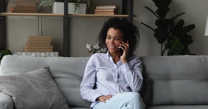 Smiling young African ethnicity woman talk on the phone. Attractive female spend day off at home sit on sofa in living room alone having pleasant remote conversation using modern tech and connection