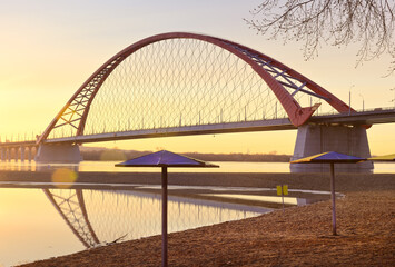 Dawn of the Bugrinsky Bridge. A deserted beach in spring, an arched modern road bridge over the Ob River in golden light. Novosibirsk, Siberia, Russia