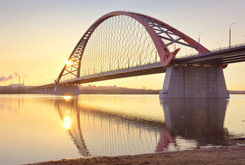 Dawn of the Bugrinsky Bridge. Arched modern automobile bridge over the Ob River in the golden morning light, the rising sun, reflections in the water. Novosibirsk, Siberia, Russia