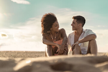 Couple of Gen Z young interracial young people in love talking sitting on the sand on the beach at sunset