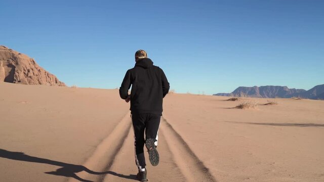 Cinematic slow motion shot of a young white man tourist with streetwear cap and boots running on a clear and beautiful sunny day with sunlight and blue sky in the Wadi Rum desert in Jordan