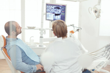 Obraz premium Senior stomatologist woman discussing with sick man about healdcare treatment looking at tooth treatment during stomatology hospital office. Orthodontic doctor examining dental x-ray