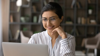 Close up smiling Indian businesswoman in glasses looking at laptop screen, working on project, reading good news in email, positive young woman using computer, student studying online at home office