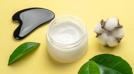 A jar of cream, green leaves and cotton on a yellow background. Concept of skin care and choice of cosmetics with a natural organic composition