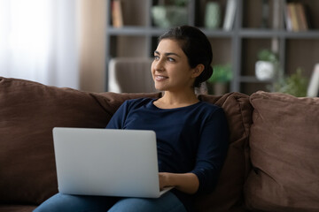 Smiling dreamy Indian woman distracted from laptop, looking to aside, sitting on couch at home, positive female visualizing future, dreaming about new career opportunity, chatting with boyfriend