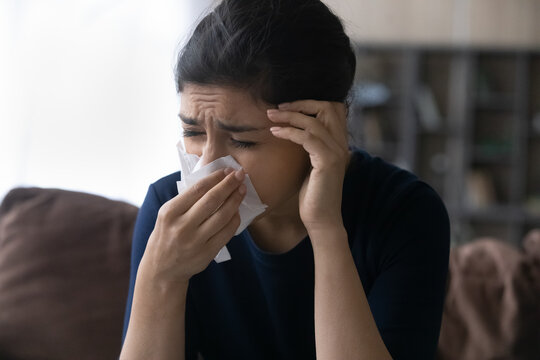 Close up unhappy sick Indian woman blowing nose, sitting on couch at home, catch cold, unhealthy young female feeling unwell, holding tissue, suffering from flu or virus, respiratory disease