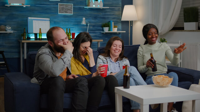 Happy multiethnic friends smiling together while sitting in frot of television looking at funny comedy movie. Group of mixed race people relaxing on couch late at night in living room