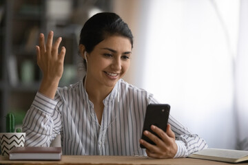Obraz premium Close up smiling Indian woman in earphones chatting on smartphone, using webcam, making video call to relatives or friends, sitting at desk, businesswoman freelancer involved in internet conference