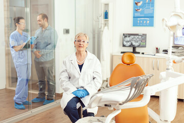 Obraz premium Portrait of senior dentist woman sitting on chair in stomatological hospital office. In background dentist assistant talking with sick patient for dental treatment during stomatology consultation