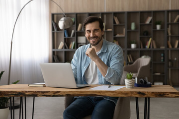 Fototapeta na wymiar Portrait of smiling successful businessman sitting at modern wooden work desk with laptop, looking at camera, happy young man student or freelancer working on project online, posing for photo