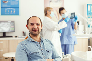 Obraz premium Man patient looking into camera waiting for radiography results sitting on dental chair in hospital stomatology clinic. In background dentists team looking at tooth x-ray preparing for surgery