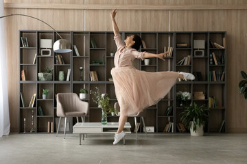 Overjoyed Indian woman wearing dress dancing, jumping in modern living room interior alone, having...