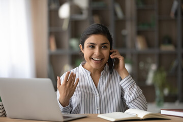 Overjoyed Indian woman talking on phone, hearing good news, sitting at desk with laptop, chatting...