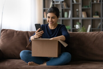 Smiling Indian young woman using smartphone, satisfied customer unpacking parcel box at home, checking internet store order, blogger shooting unboxing video, good quick delivery service concept