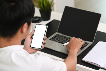Back view of young man using mobile phone and working with laptop computer at office.