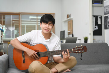Happy young asian man smiling and sitting with his guitar on sofa at home.