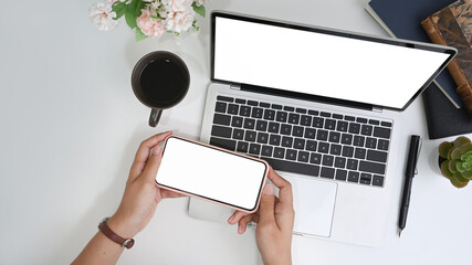 Above view of woman holding horizontal mobile phone with blank screen over laptop computer at...