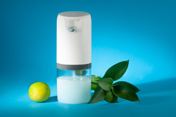 Soap dispenser automatic. Close-up. Nearby there is a lime and a green branch. Blue background.